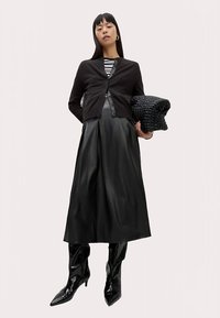 Schwarze Strickjacke über einem gestreiften Oberteil, kombiniert mit einem schwarzen, plissierten Rock aus Kunstleder und kniehohen schwarzen Stiefeln; hält eine strukturierte schwarze Tasche.