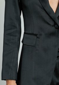 Blazer a righe nere con una texture liscia, dotato di una piccola tasca a patta e rever a punta. Il tessuto ha una leggera lucentezza.