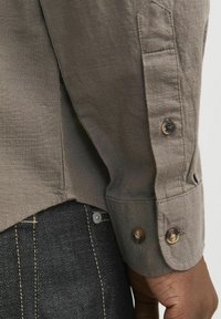 Long-sleeve, olive green shirt made of a textured fabric, featuring buttoned cuffs and a side split hem, worn over dark denim jeans.