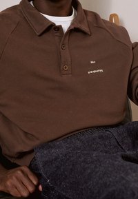 Polo shirt marrone con colletto, caratterizzato da due bottoni marroni e testo bianco. Realizzato in tessuto testurizzato, indossato sopra jeans in denim scuri.