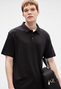 Camisa polo preta de mangas curtas com colarinho e um fecho de três botões; apresenta um tecido macio e é acompanhada de uma bolsa preta com ferragens prateadas.