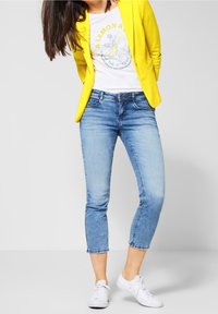 Světle modré džíny s přiléhavým střihem a zkrácenou délkou, doplněné žlutým sako a b ílým tričkem s potiskem. Bílé tenisky dotvářejí celkový vzhled.