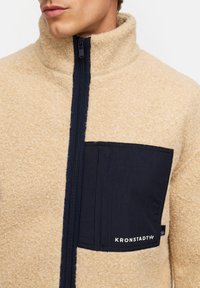 Beige fleecetröja med hög krage, med en mörk marinblå bröstficka med en logotyp och synlig dragkedja. Mjuk textur.