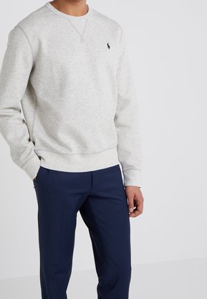 Man draagt lichtgrijze trui met klein zwart paardenlogo en donkerblauwe broek, staand met handen in de zakken.