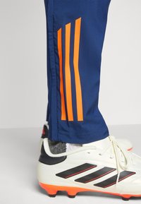 Pantaloni sportivi navy con design a tre strisce arancioni, realizzati in tessuto liscio. Presentano scarpini da calcio bianchi e neri con dettagli arancioni.