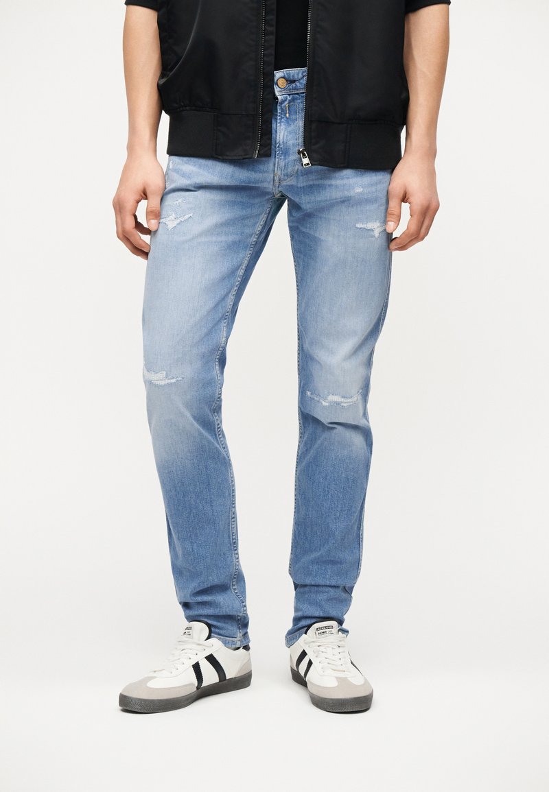 Άνδρας που φοράει ανοιχτό μπλε τζιν με φθορές, λευκά αθλητικά παπούτσια με μαύρες ρίγες και μαύρο σακάκι, στέκεται μπροστά σε λευκό φόντο.