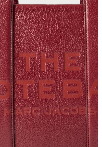 Rde teksturirano usnje s stisnjenimi velikimi črkami "THE" in manjšim napisom "MARC JACOBS" spodaj.