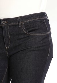 Mörkblå denimjeans har en standard femficksdesign, sydda sömmar och en knappstängning i midjan. Slät textur och regular fit.