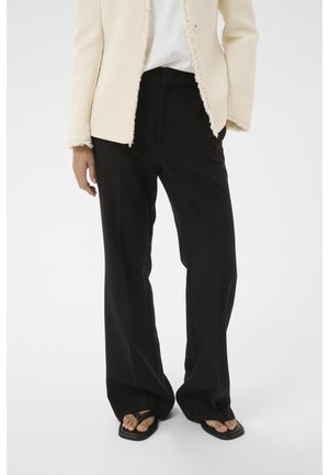 Personne portant un pantalon noir à jambes larges, une veste texturée crème, un haut blanc et des sandales noires à bout carré avec brides pour les orteils.