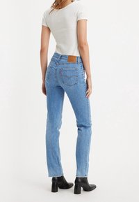 Jeans de mezclilla azul claro con un ajuste ajustado, que presentan un parche de cuero marrón con marca en la parte posterior, combinados con una blusa blanca de manga corta y tacones negros.