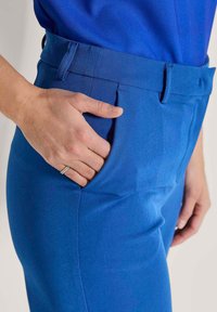 Blauwe op maat gemaakte broek met een zachte textuur, met zijzakken en een riemlus, weergegeven naast een hand die in de zak rust.