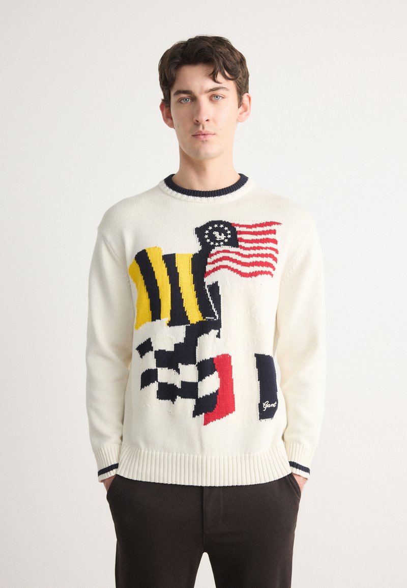 Pull en tricot crème avec de grands motifs de drapeaux multicolores, col et poignets côtelés navy, et un petit logo en bas.