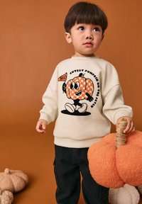 Cremefarbener Sweatshirt mit einem cartoonhaften Kürbis-Design in karierter Optik und dem Text "Süßester Kürbis im Feld", kombiniert mit schwarzen Hosen.
