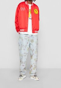 Bomber jacket in raso rosso con righe bianche, t-shirt grafica, pantaloni in denim chiaro con un motivo colorato e sneakers bianche stampate.