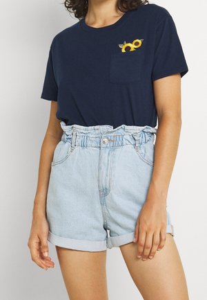 Kvinde iført en mørkeblå t-shirt med solsikker på lommen og lyseblå højtaljede denimshorts med opbrettede kanter.