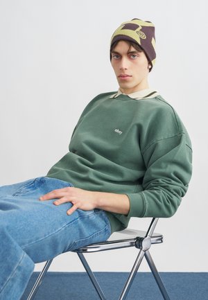 Νεαρός που φοράει καμουφλαρισμένο σκουφάκι, πράσινο φούτερ Obey και μπλε τζιν, καθισμένος χαλαρά σε μεταλλική καρέκλα μπροστά σε απλό φόντο.