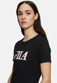 T-shirt en coton noir avec un col rond, arborant un logo "FILA" blanc et rouge sur le devant, texture lisse et manches courtes.