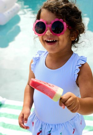 Lächelndes Kleinkind mit rosa, blumenförmiger Sonnenbrille und blau gestreiftem Badeanzug hält ein in Form einer Melone geformtes Eis am Stiel neben einem Pool.