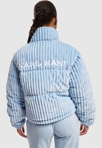 Světle modrá péřová bunda s žebrovanou strukturou, vysokým límcem a vyšitým logem "KARL KANI" na zadní straně. Měkká, plyšová tkanina.