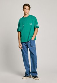 Camiseta de algodón verde con mangas cortas, con gráficos blancos, combinada con jeans de mezclilla azul claro y zapatillas negras con suelas blancas.