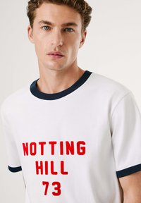 Λευκό βαμβακερό T-shirt με κόκκινο κείμενο "NOTTING HILL 73", ολοκληρωμένο με ναυτικό μπλε ριμπ τριμ στον λαιμό και στα μανίκια. Σχεδίαση κανονικής εφαρμογής.