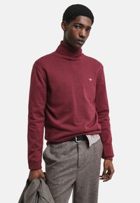 Maglione a collo alto bordeaux in morbido knit con colletto a costine. Abbinato a pantaloni grigi a fantasia. Il modello tiene una giacca su un braccio.