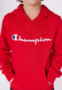 Sudadera roja hecha de algodón suave, con un bolsillo delantero, mangas acanaladas y un logo bordado en blanco de "Champion" en el pecho.