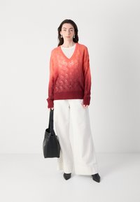 Pull en tricot avec un dégradé d'orange à rouge, orné d'un motif géométrique, à encolure en V et à manches longues. Associé à un pantalon large blanc et un sac noir.