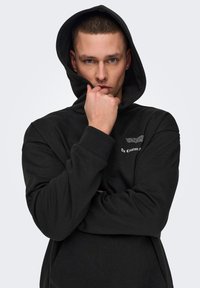 Sudadera negra con capucha que presenta un bolsillo frontal tipo canguro, puños acanalados y un estampado gráfico en el pecho. Textura suave y corte casual.