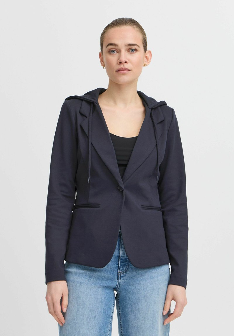 Blazer blu navy con cappuccio, realizzato in tessuto morbido, con due tasche frontali e una vestibilità sagomata, indossato sopra a una maglia nera e jeans chiari.