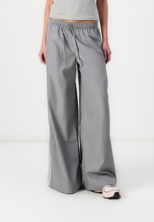 Person trägt graue, weit geschnittene Hosen mit Kordelzug in der Taille und hellrosa Sneakers, steht vor einem schlichten weißen Hintergrund.