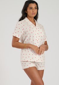 Ensemble de pyjama à manches courtes en tissu crème, avec un motif en forme de cœur rouge. Comprend un haut à boutons avec poches et un short assorti.