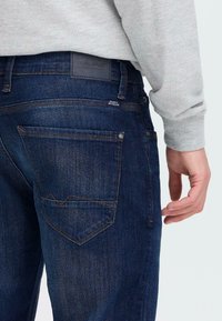 Primer plano de una persona con jeans de mezclilla azul oscuro y una camiseta de manga larga gris claro, mostrando el bolsillo trasero y la mano relajada.