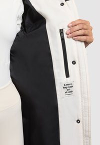 Balta pūkinė striukė su juodu pamušalu, užtrauktuko kišenė ir etiketė su tekstu: „Šilta apkabinimas iš audinio.“