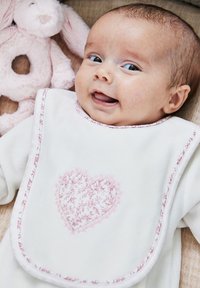 Valkoinen essu, jossa on vaaleanpunainen kukkakuviollinen sydän, vaaleanpunaisilla reunuksilla, jota vauva käyttää. Essu on pehmeän vaaleanpunaisen pupulelun vieressä beige taustalla.