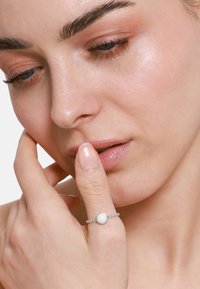 Anello d'argento con una pietra bianca centrale, accentuato da piccole pietre trasparenti lungo la fascia, indossato su un dito vicino alla bocca.