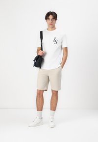 Witte t-shirt met "LS" in grijs, gecombineerd met beige shorts. Het model draagt witte sneakers en heeft een zwarte schoudertas. Slim fit.