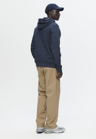 Un hoodie marine avec une poche arrière, un pantalon beige et des baskets grises, présenté de côté sur fond blanc.