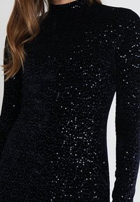 Svart långärmad klänning i plyschtyg, täckt av glittrande paljetter, med hög halsringning och figurnära design.