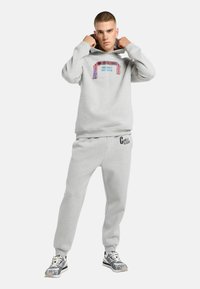 Ung mand der justerer hætten på sin lysegrå sweatshirt med farverigt Milano-design, matchende grå sweatpants og mønstrede sneakers på hvid baggrund.