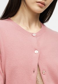 Κοντινό πλάνο γυναίκας που φοράει μαλακό ροζ πουλόβερ με κουμπιά, με μακριά σκουρόχρωμα μαλλιά μερικώς ορατά, εστιάζοντας στον λαιμό και στα κουμπιά.