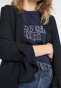 Námořnický blejzr s rozšířenými rukávy, zkombinovaný s námořnickým tričkem s bílým textem. Oblékáno s modrými džíny s vysokým pasem, které vykazují jemnou strukturu.