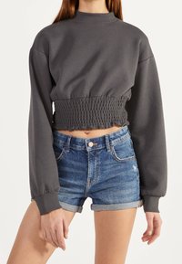 Grauer, verkürzter Sweatshirt mit langen Ärmeln und gerüschtem Bund, kombiniert mit blauen Denim-Shorts. Das Material wirkt weich und texturiert.
