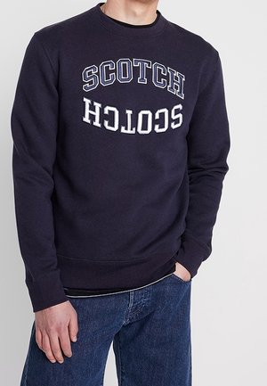 Homme portant un sweat-shirt bleu marine avec le mot « SCOTCH » imprimé deux fois, une fois normalement et une fois à l'envers, accompagné d'un jean bleu.
