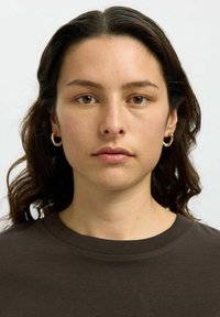 Smeđa majica s okruglim izrezom; model ima srednje dugu, valovitu tamnu kosu; nosi zlatne naušnice na prsten; neutralna pozadina.