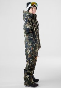 Camouflage sneeuwpak met een rits aan de voorkant, capuchon en zijdezakken. Voorzien van oranje ritsaccenten en zwarte sneeuwlaarzen. Transparante goggles op het hoofd.