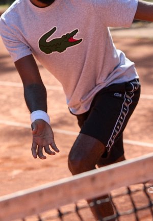 Männlicher Tennisspieler, der ein graues Shirt mit Krokodillogo und schwarze Shorts trägt und sich auf einem Sandplatz nahe dem Netz nach vorne streckt.