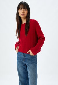 Roter gerippter Pullover mit langen Ärmeln, lockerer Passform und Rundhalsausschnitt, kombiniert mit hochtaillierten blauen Jeans. Einfaches Design, einfarbig.