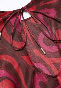 Tissu à motifs rouges et roses avec une texture lisse, présentant un détail œillet noué avec de longs cordons et des accents métalliques.