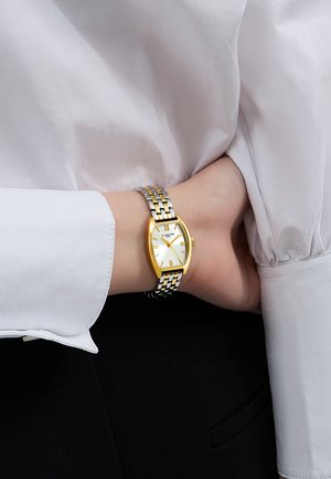 Montre en or et argent avec un boîtier courbé, un cadran blanc, des index horaires en or et un bracelet métallique à maillons. Portée sur un poignet dans une manche blanche.